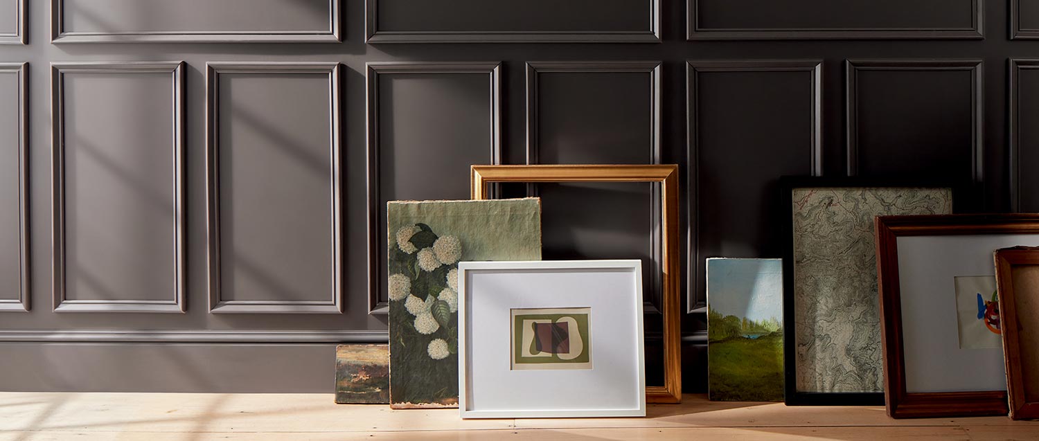 Artwork and frames arranged on a floor against a deep chocolate brown-painted wall with decorative molding.